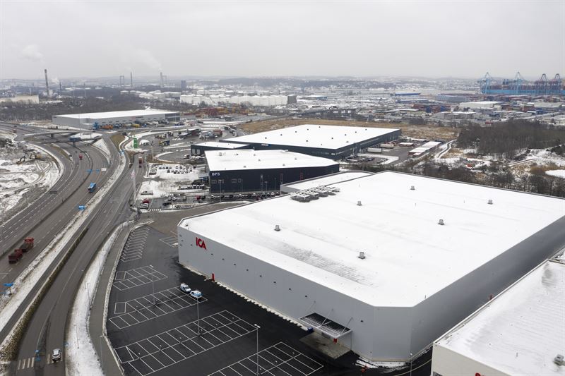 Bild tagen utifrån på ett stort gråvitt Ica-lager med kringliggande parkering och motorväg i vintrigt väder.